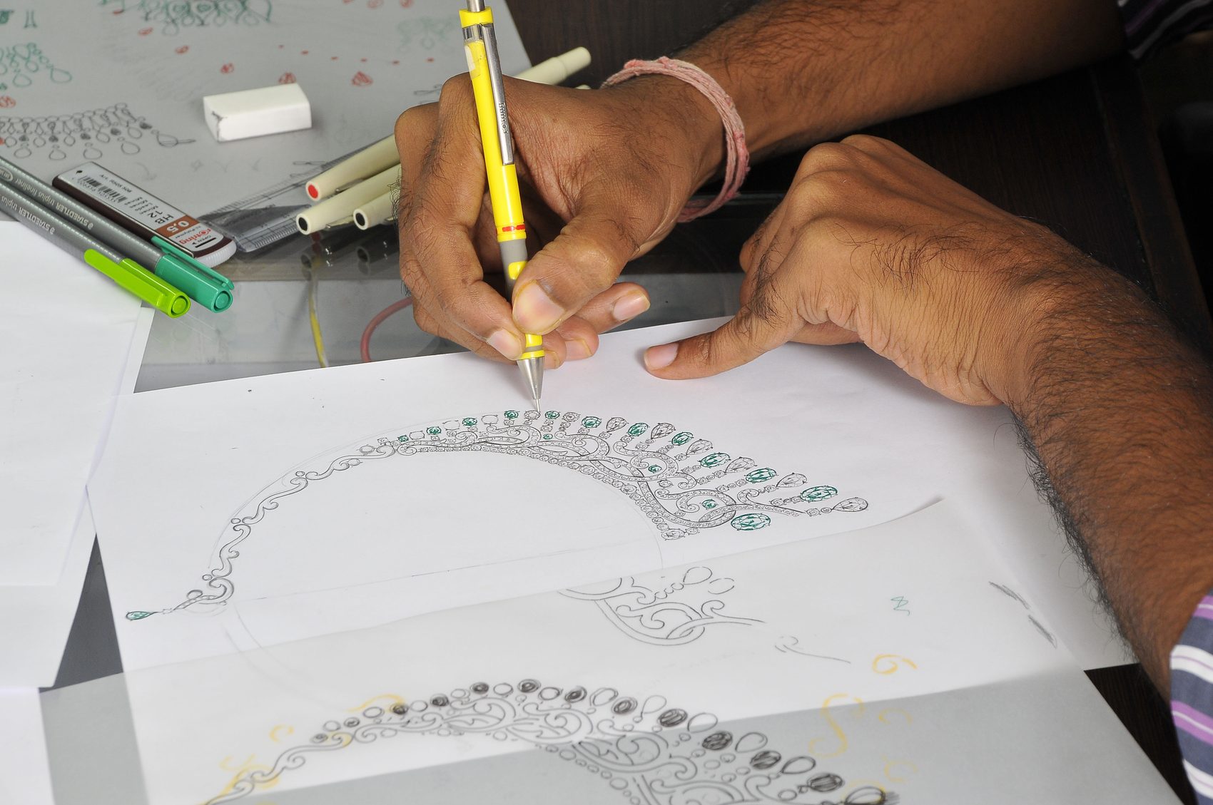 A designer's hands sketching a jewellery design in pencil at the studio bench